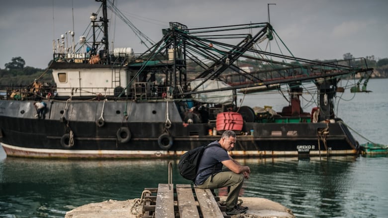 A man sits at the water's edge, looking tired and forlorn, while a massive fishing vessels sits on the water behind him. 