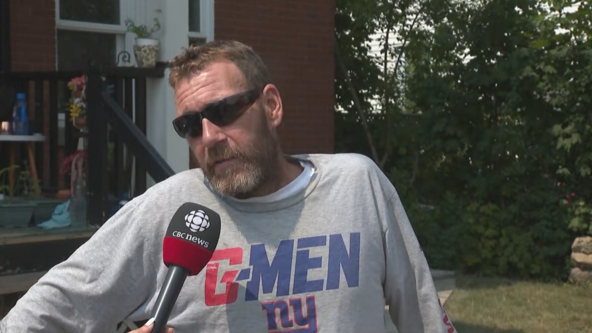 A middle-aged man in sunglasses and a sweatshirt, visible from the stomach up, speaks into a reporter's microphone outside a brick house on a sunny summer day.