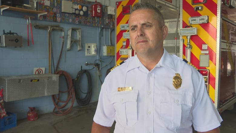 A white man with greyish hair stands in front of the back end of a fire truck. He's wearing a white, short sleeved uniform shirt.