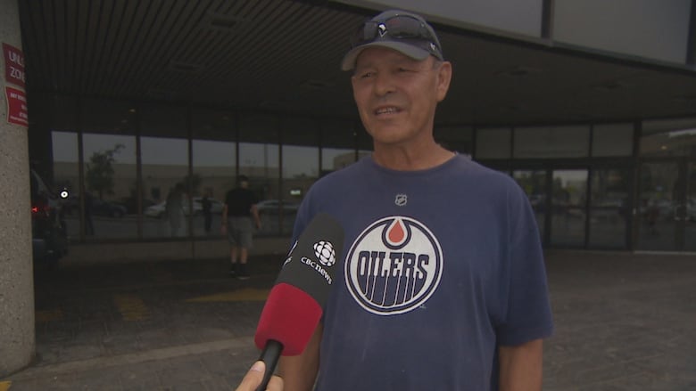 A man in a baseball cap and T-shift speaks into a mic in front of a glass building.