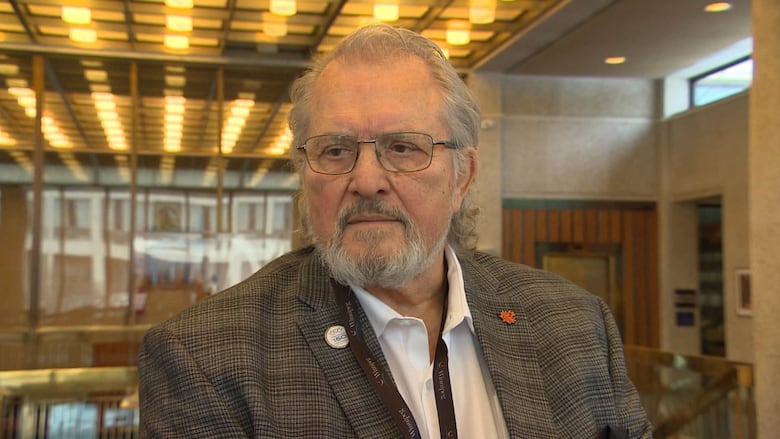 Man with white hair and eyeglasses wears a grey tweed suit jacket.