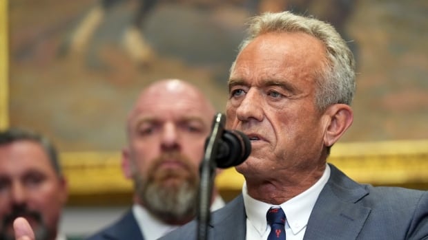 A man speaks into a microphone. Other people are visible standing behind him. 
