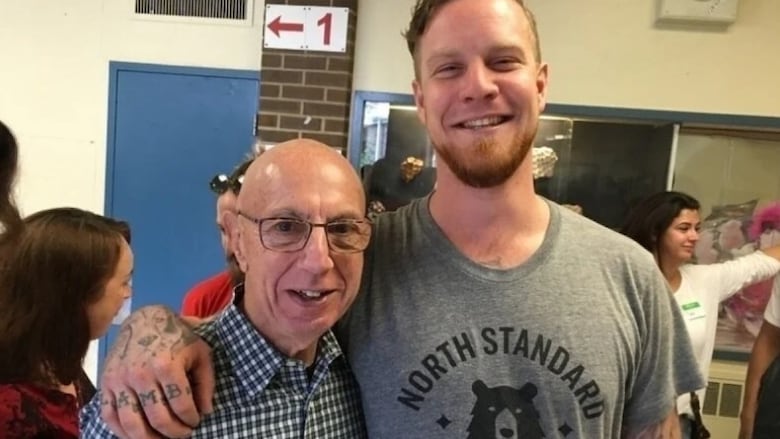 A bald, senior man in a checkered shirt stands arm in arm with a student inside a school with other students passing by in the background