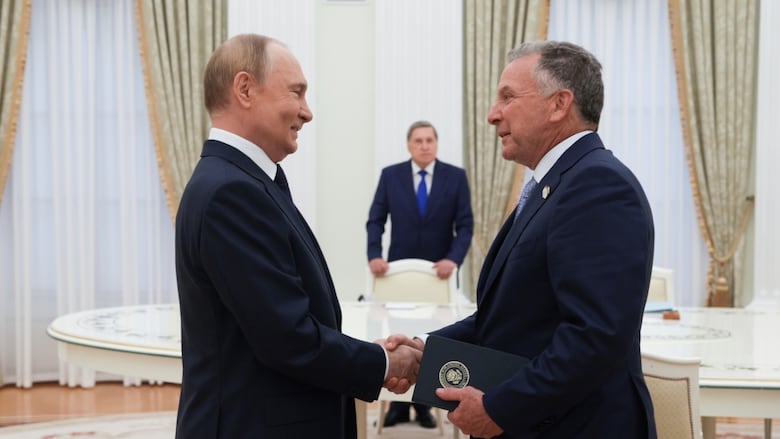 Two older clean shaven men wearing suit and tie shake hands and smile while standing in an ornate room.