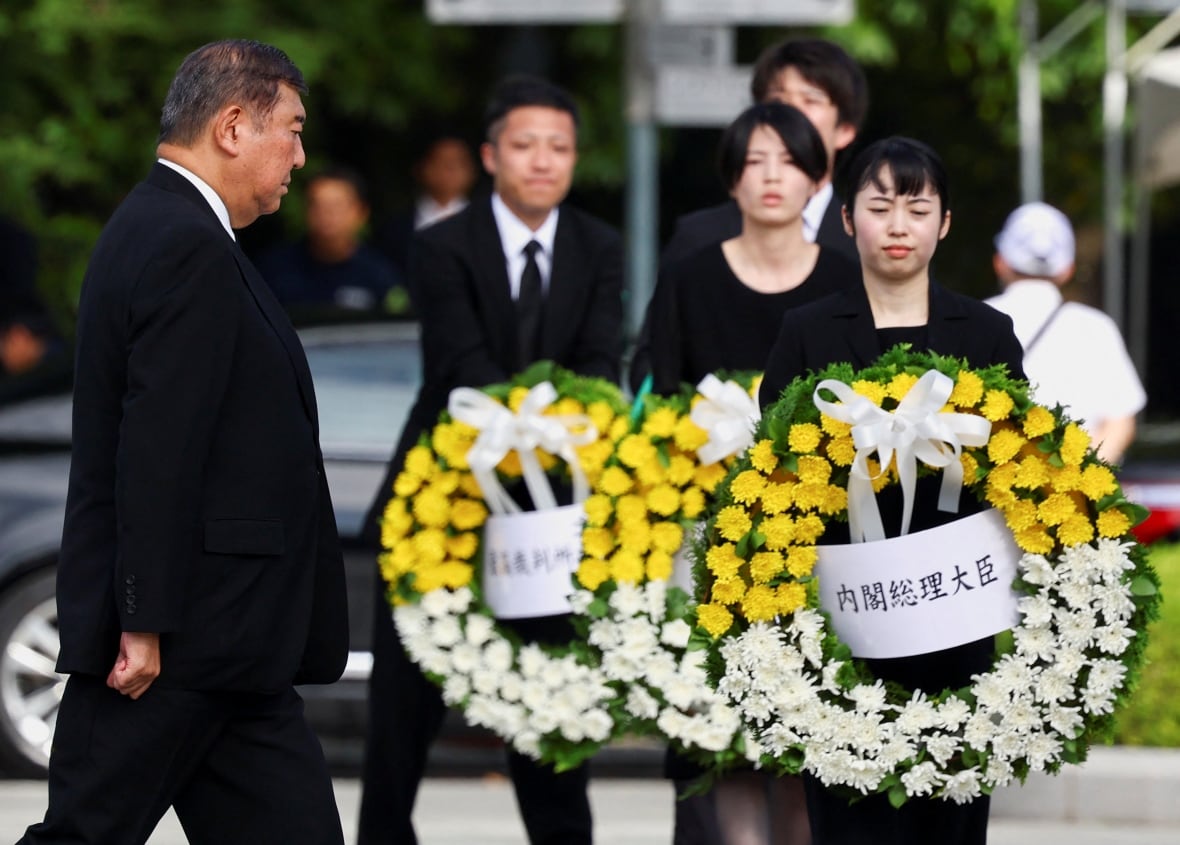 An older Asian man wears a dark suit near Asian women carrying a large flower wreaths.