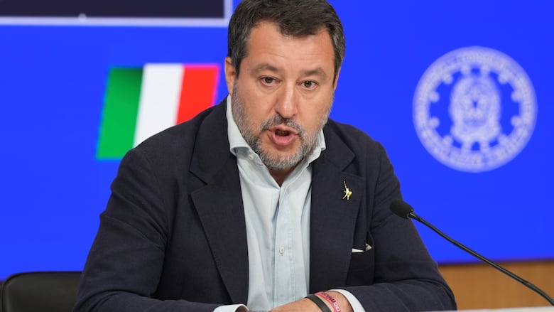 A bearded man wearing a blazer and collared shirt speaks while seated.