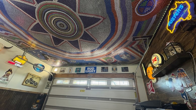 A picture of a garage that with beer signs on the walls. Its ceiling has a mosaic made up of of thousands beer caps. It's multi-coloured, and features different designs.