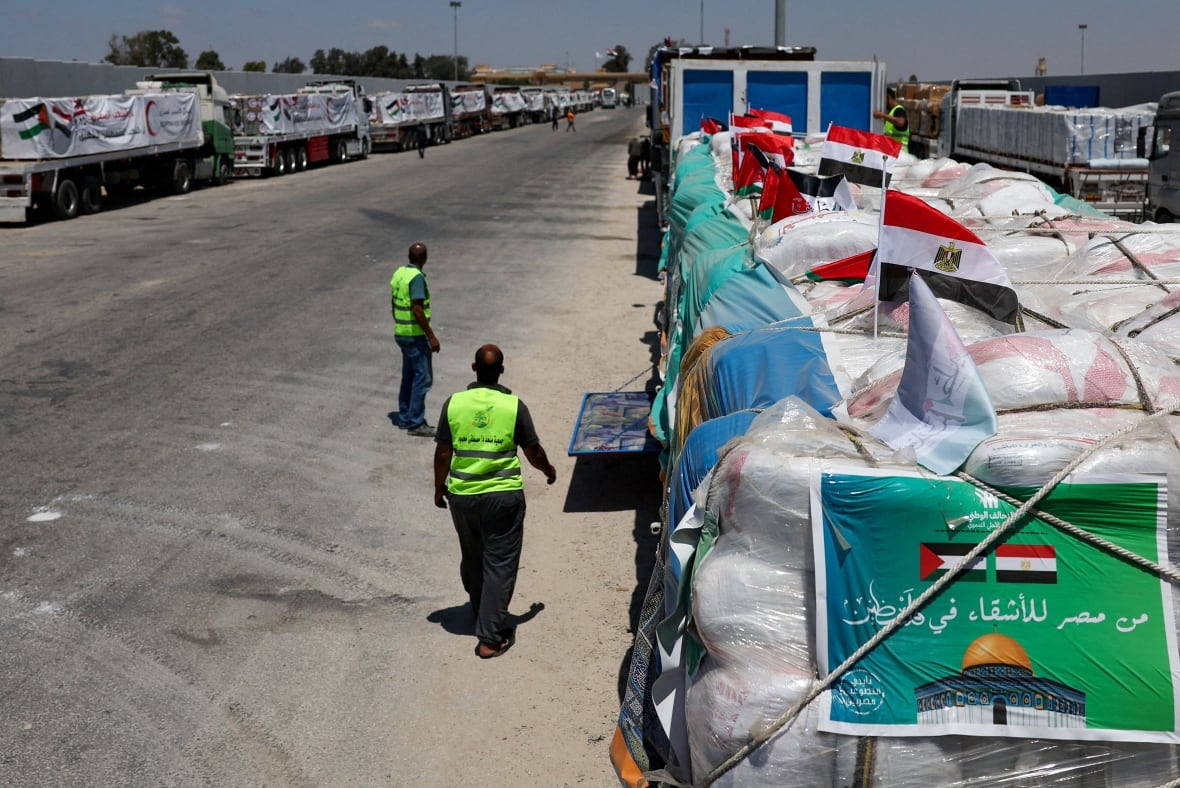 People walk next to trucks carrying humanitarian aid on a hot day.