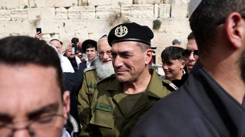 A man in military uniform, including a black beret, is surrounded by security and reporters.