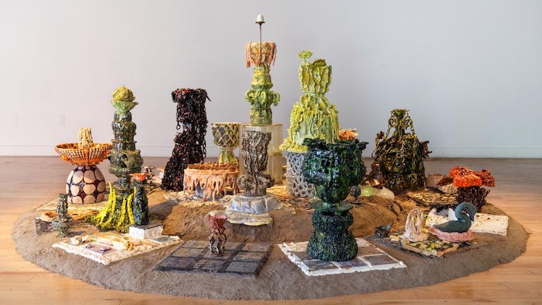 In a large exhibition space, an assortment of ceramic vessels resembling organic shapes sit on top of a circular mound of sand.