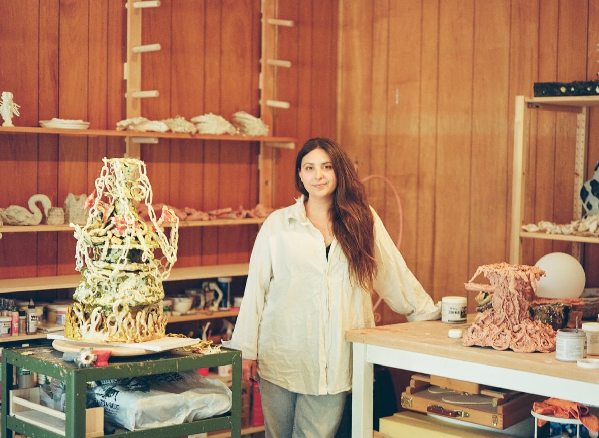 A woman with long dark hair, a white buttoned shirt and jeans leans on a table holding a sculpture. Another sculpture stands on a table nearby.