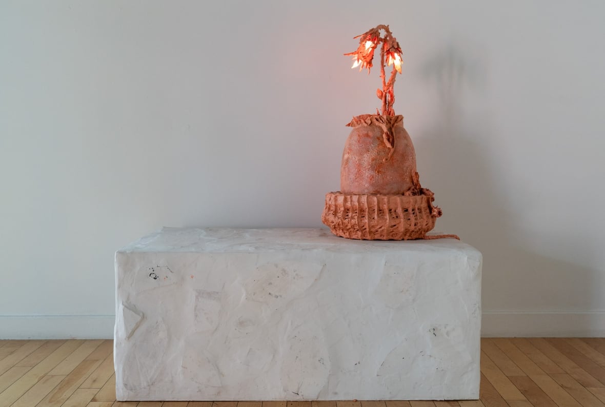 On top of a white platform sits a ceramic sculpture that looks like an egg with a plant sprouting from its top. The plant holds three red lightbulbs. 