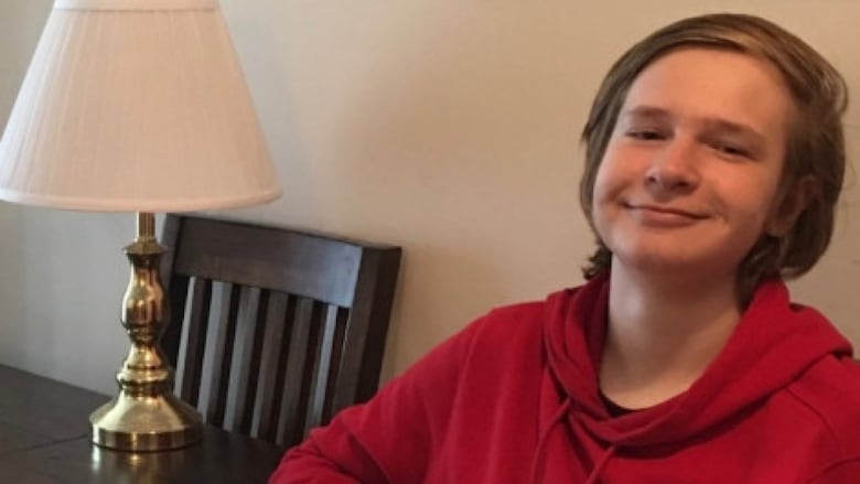 A teenage boy with blonde/light-brown hair wearing a red hoodie sits at a dining room table, smiling.
