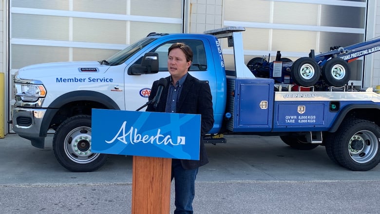 A man in a blue suit jacket and jeans stands in front of a blue podium with a tow truck and an industrial garage in the background.