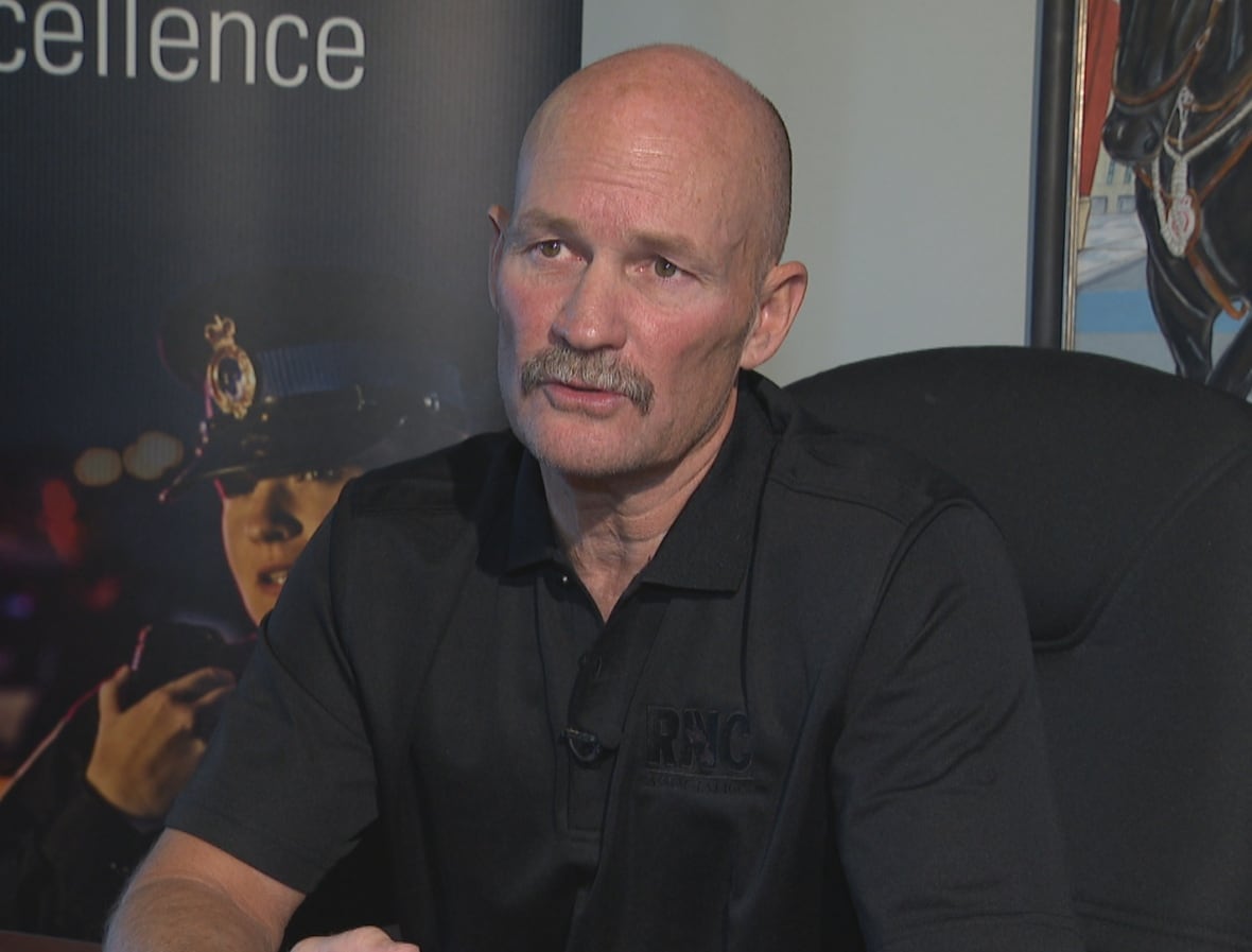 A bald, middle aged man sits in a black chair in a board room with a painting of a police officer on a horse behind him.