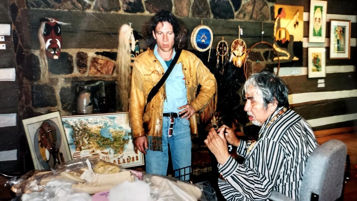 A man in a fringe leather jacket with long hair stands before an old man in a chair. There is artwork around them.