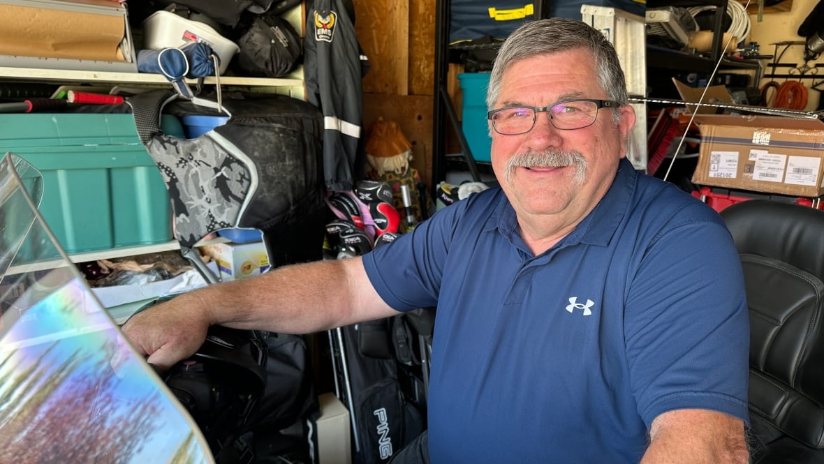 Un hombre blanco mayor, con cabello gris corto y un bigote gris espesado, lleva vasos y una camisa de armadura azul marino. Está sentado en una motocicleta estacada dentro del garaje de su casa, que está lleno de cajas, bolsas y varios otros artículos.