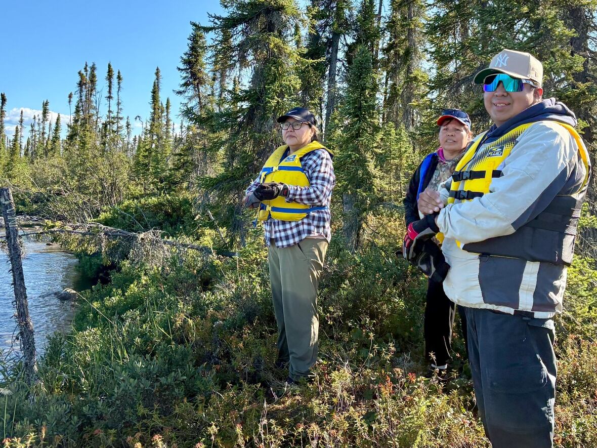 Three people wearing life jackets stand at the water's edge. 