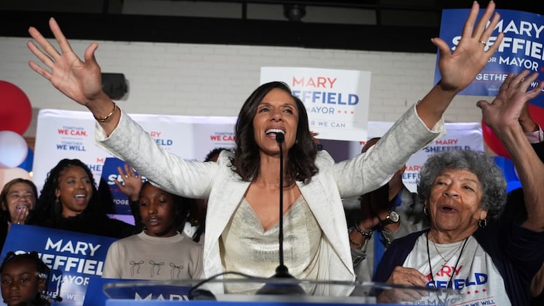 Mary standing at a podium, arms raised in victory, with supporters behind her holding signs.