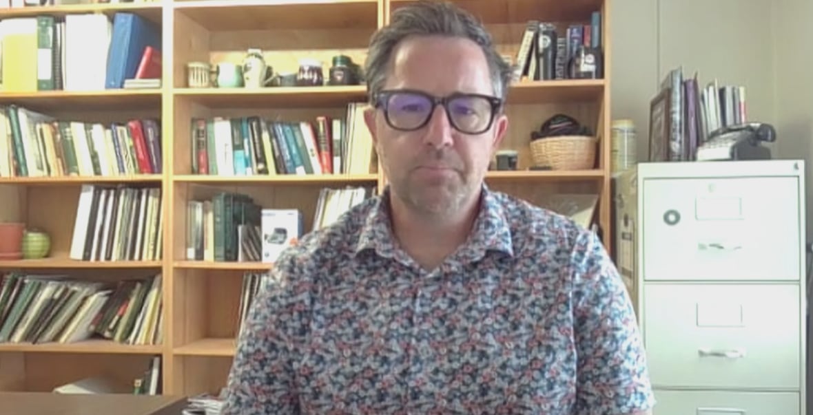 A man in a collared shirt, a bookshelf and filing cabinet is behind him.