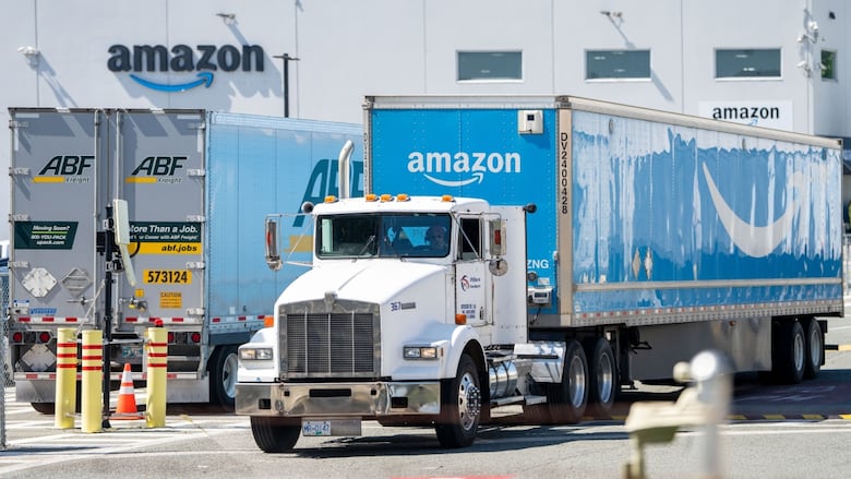 A large truck with a blue container reading 'Amazon' on it next to a large warehouse with the same symbol.