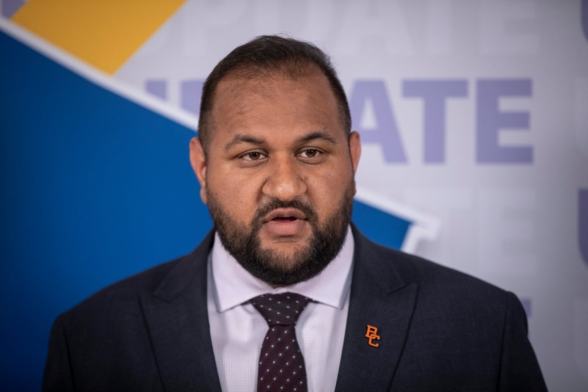 A South Asian man wearing a suit speaks in front of a screen.
