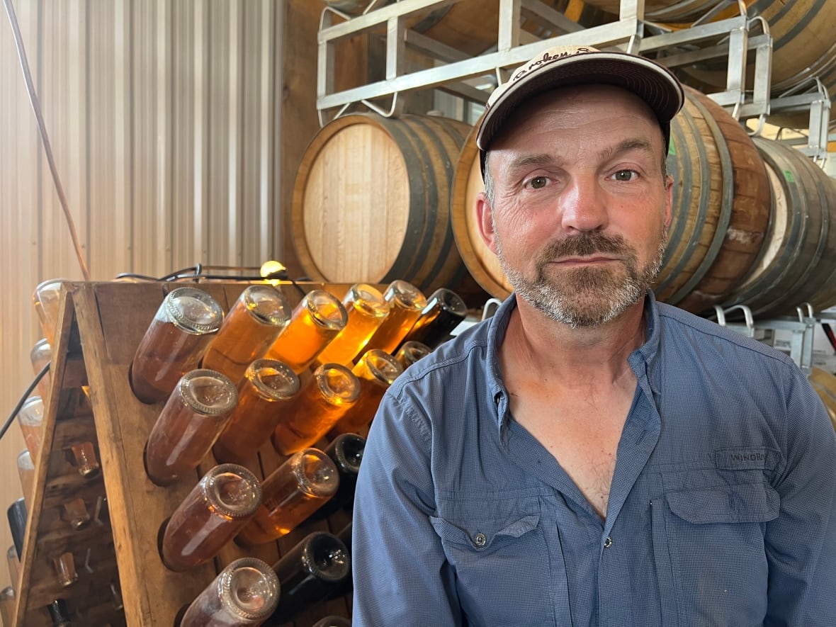 A man in a blue shirt and cap sits in front of barrels and bottles of wine.
