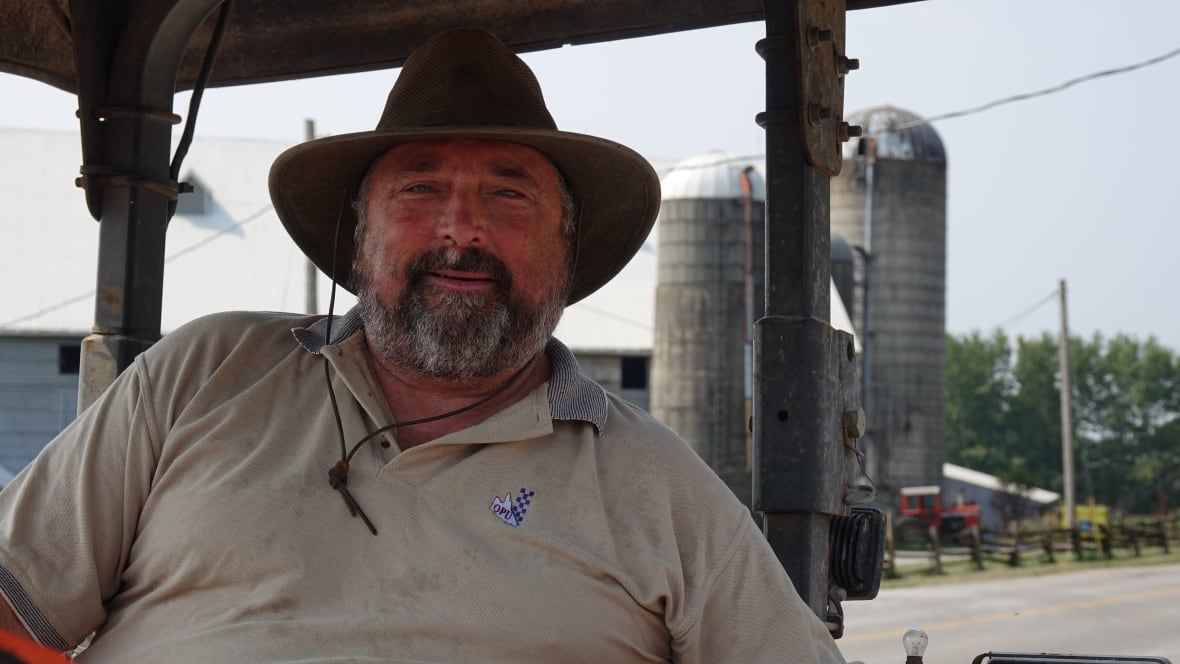 A man wearing a wide-brimmed hat and a beige polo shirt sits in a tractor in front of a barn and silos.