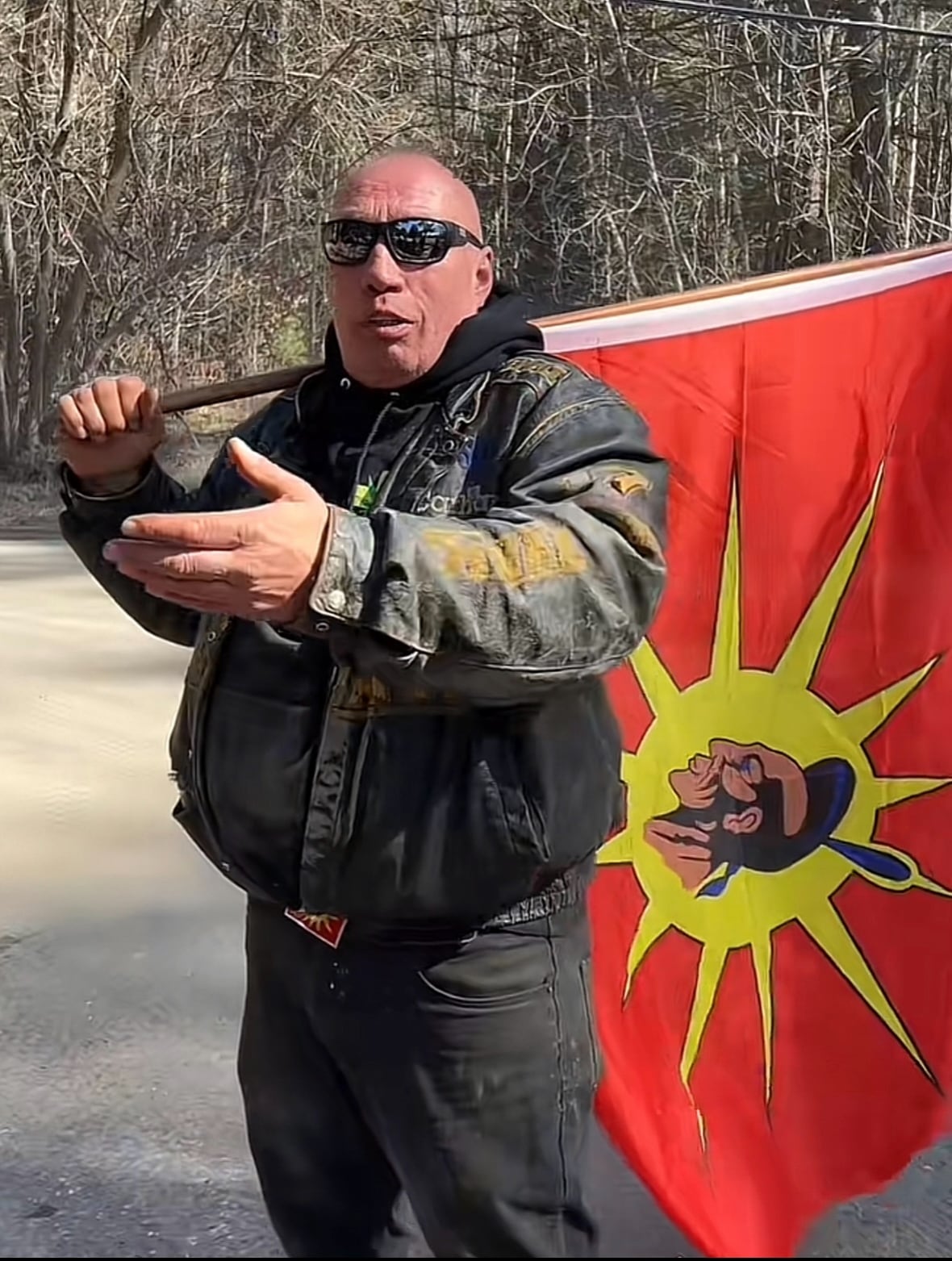 A large man with sunglasses and close-cropped hair stands looking at the camera. He is holding a flag pole with a flag over his shoulder. The flag has a red background with a yellow sunburst in the middle that surrounds the head of a warrior.