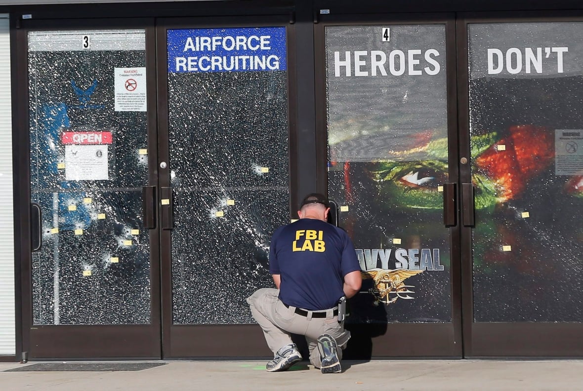 A person with their back to the camera wearing a tshirt that says FBI Lab kneels down in front of a glass door that has been shattered.