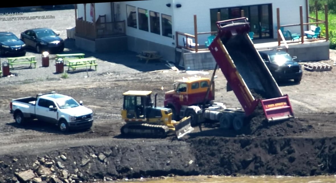 A dump truck with a load of blacksoil dumps it along the shore of a lake. Behidn the dump truck is the back of a building.
