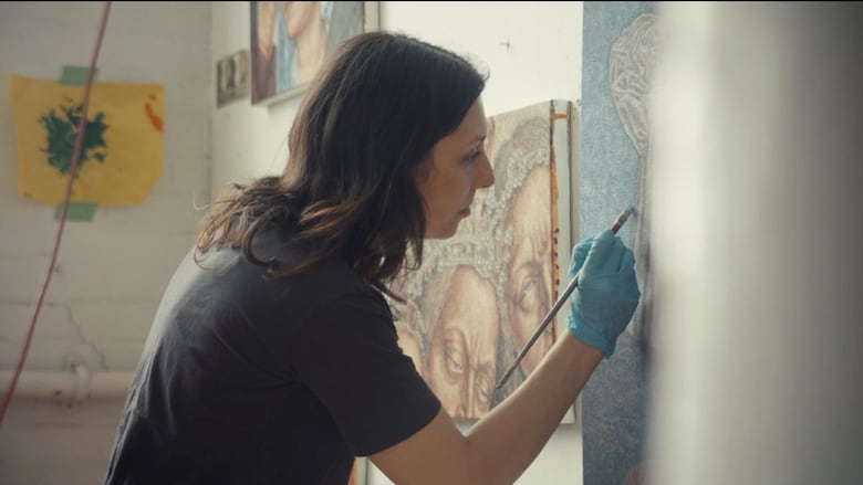 A woman with dark hair wearing a black t-shirt and blue protective gloves dabs at a canvas with a paintbrush.