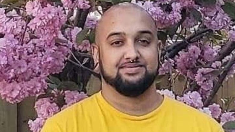 Photo shows man with beard smiling in front of pink flowers and wearing a yellow shirt. 