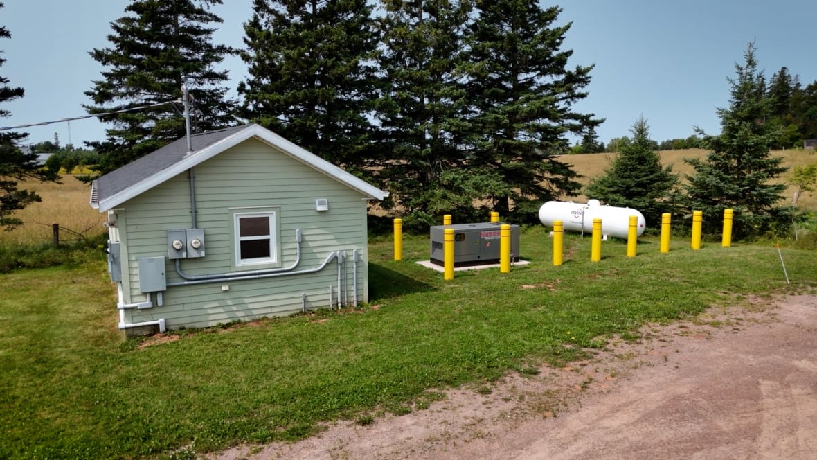 Utility shed with mounted boxes and pipes, a propane tank labeled "Irving," and yellow bollards in a grassy, tree-lined area