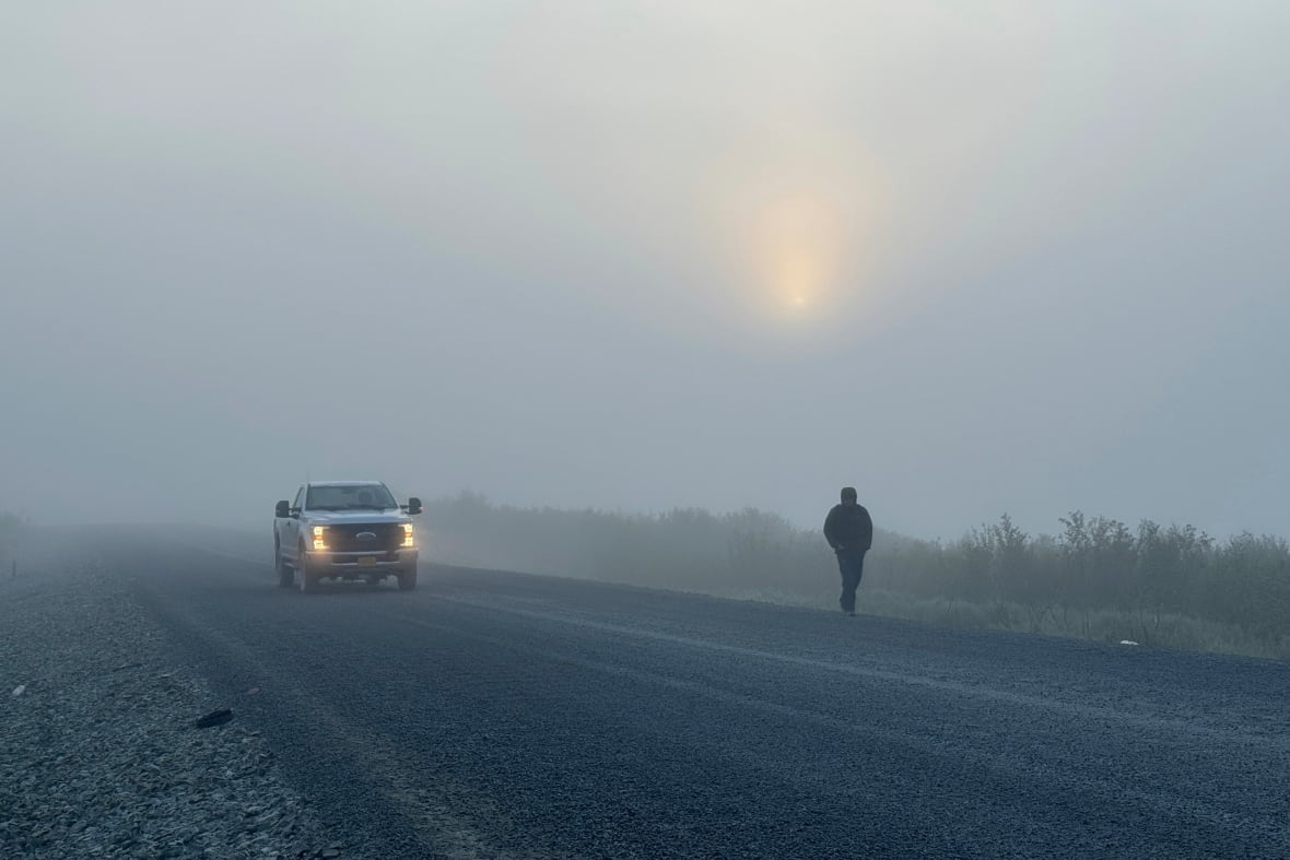 A person walks along a road in the fog as a vehicle passes.