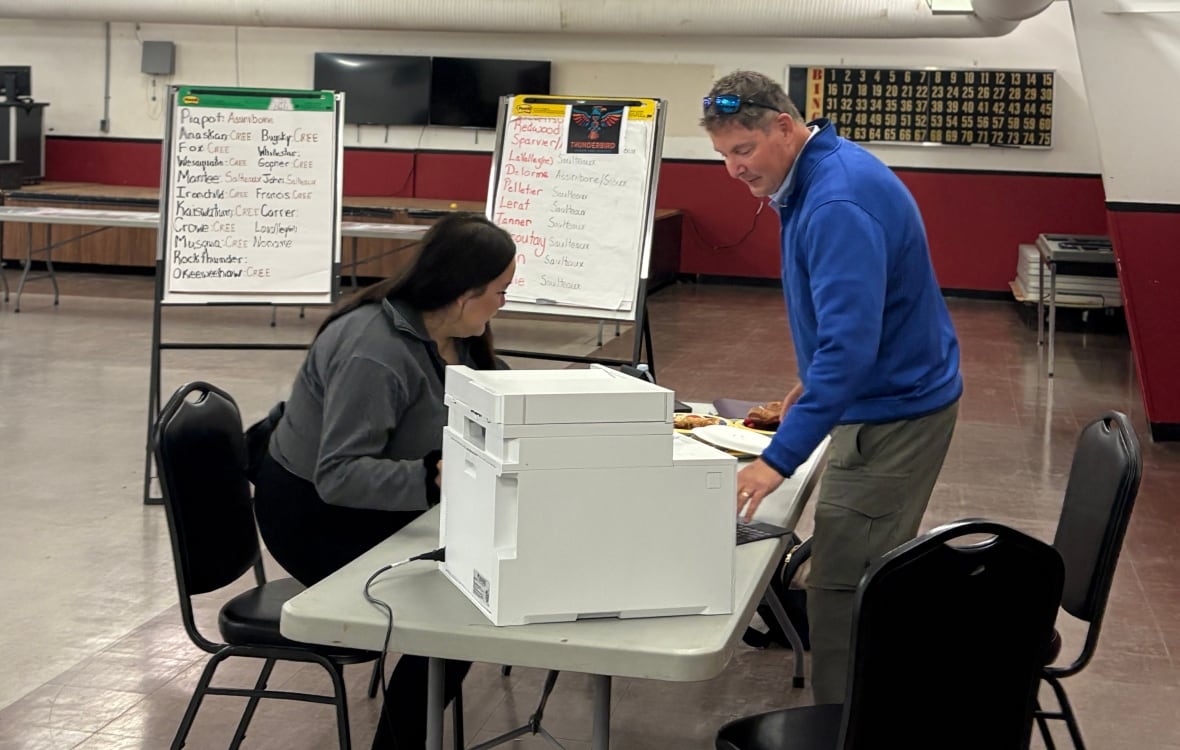 Man in right pull over sweater and short hair sits over a table with a printer, while a women with long hair sits and waits for what has been printed.