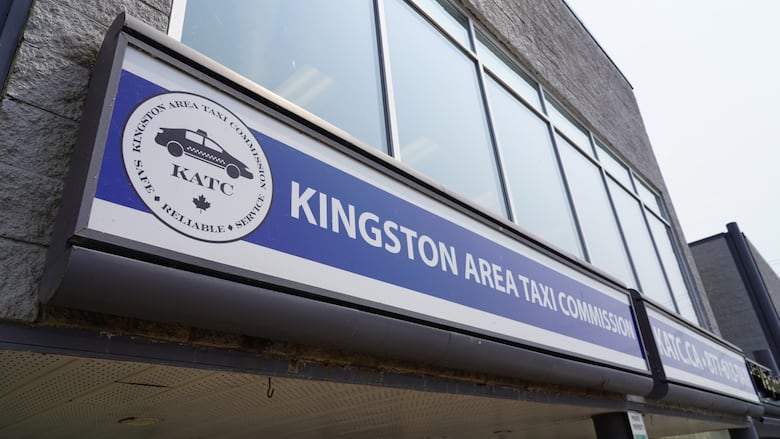 A blue sign outside a modern, building with a row of glass windows reads "Kingston Area Taxi Commission" with a logo of a cab shown beside the words.