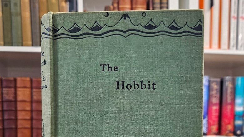 A copy of the Hobbit stands atop of other books. It is green, with the image of a dragon and mountains on the front. 