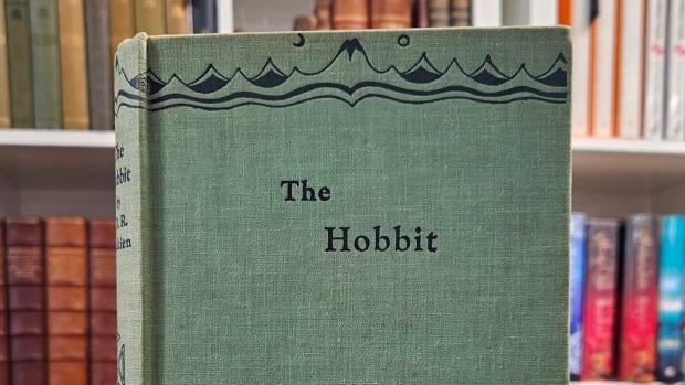 A copy of the Hobbit stands atop of other books. It is green, with the image of a dragon and mountains on the front. 