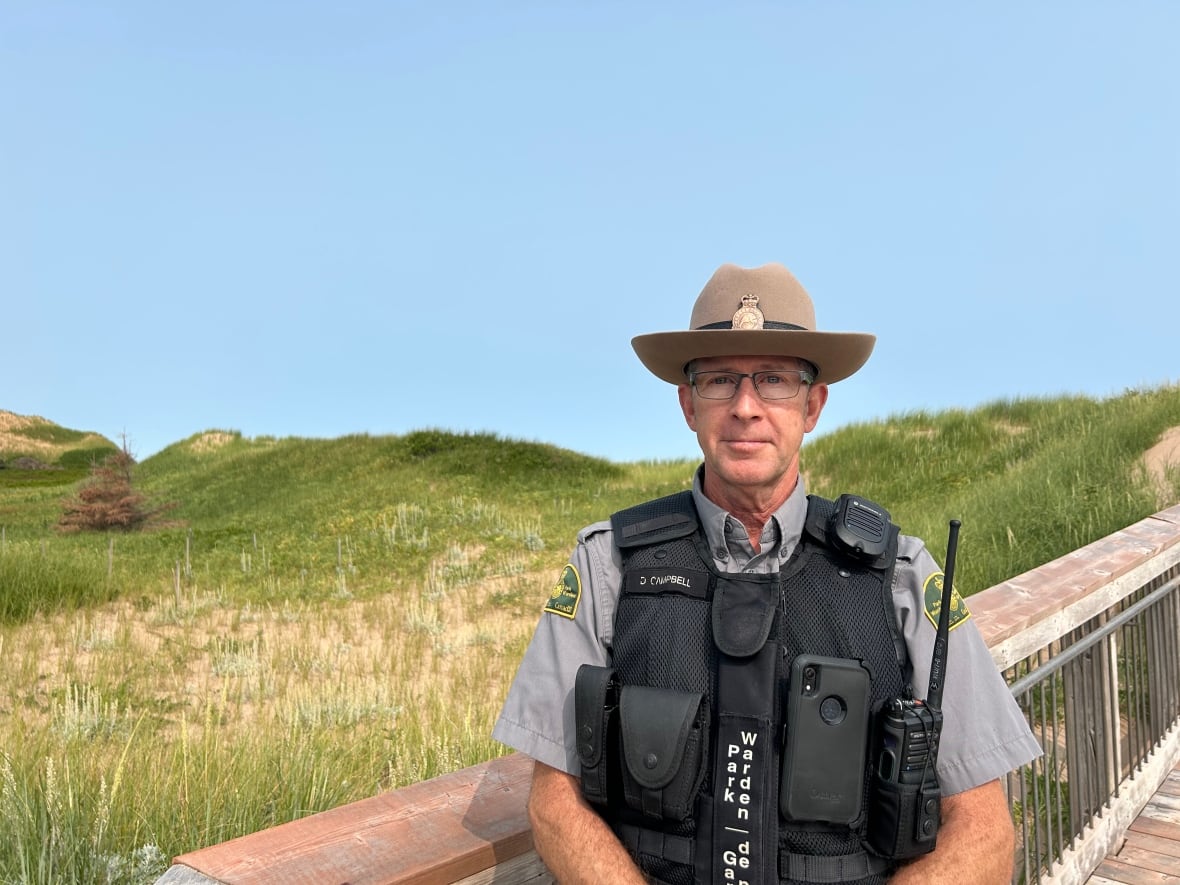 A park warden wearing a vest and hat stands on a boardwalk, sand dunes can be seen behind him.
