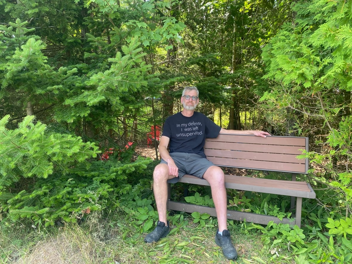 A man in glasses, shorts and t-shirt sits on a park bench in front of a wooded area.