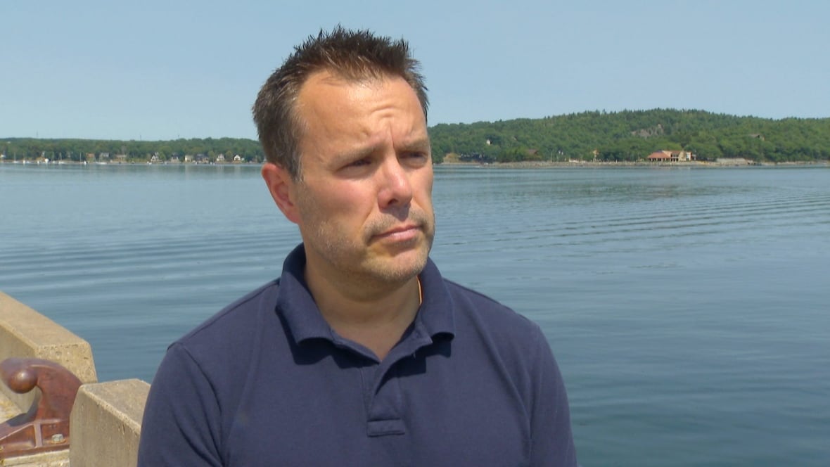 A white man with short brown hair wearing a navy polo stands outside on a jetty with water behind him, and green trees on a distant shore