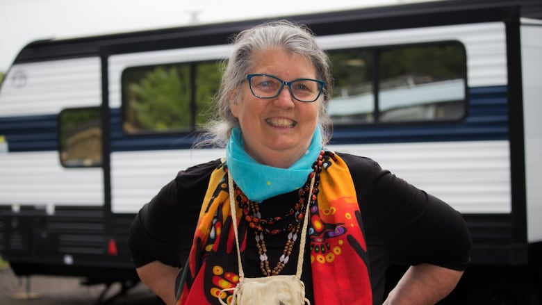 A woman in a black long sleeve shirt with an orange scarf stands smiling in front of a camping trailer