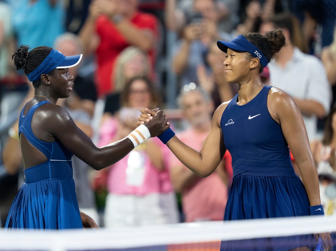 Two young women wearing blue tennis dresses and visors clasp hands on a tennis court. 