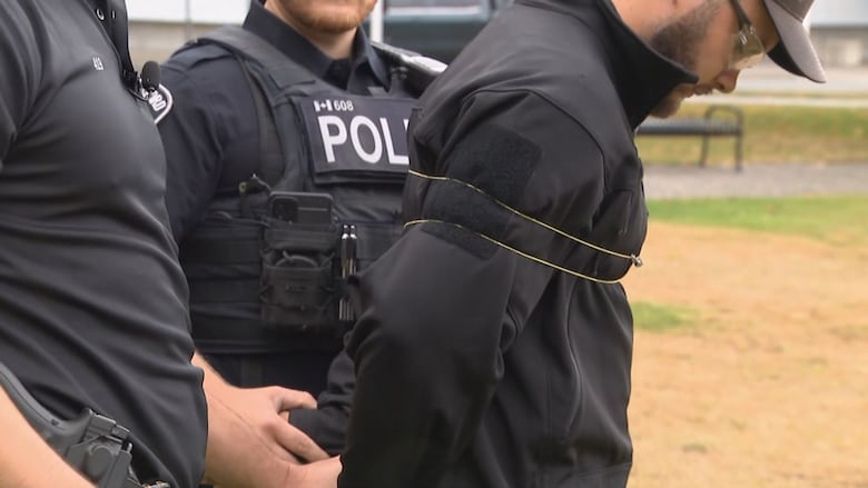 A man is restrained by a band around his upper body with two men wearing vests that say, "Police," on the front of the vest behind him.
