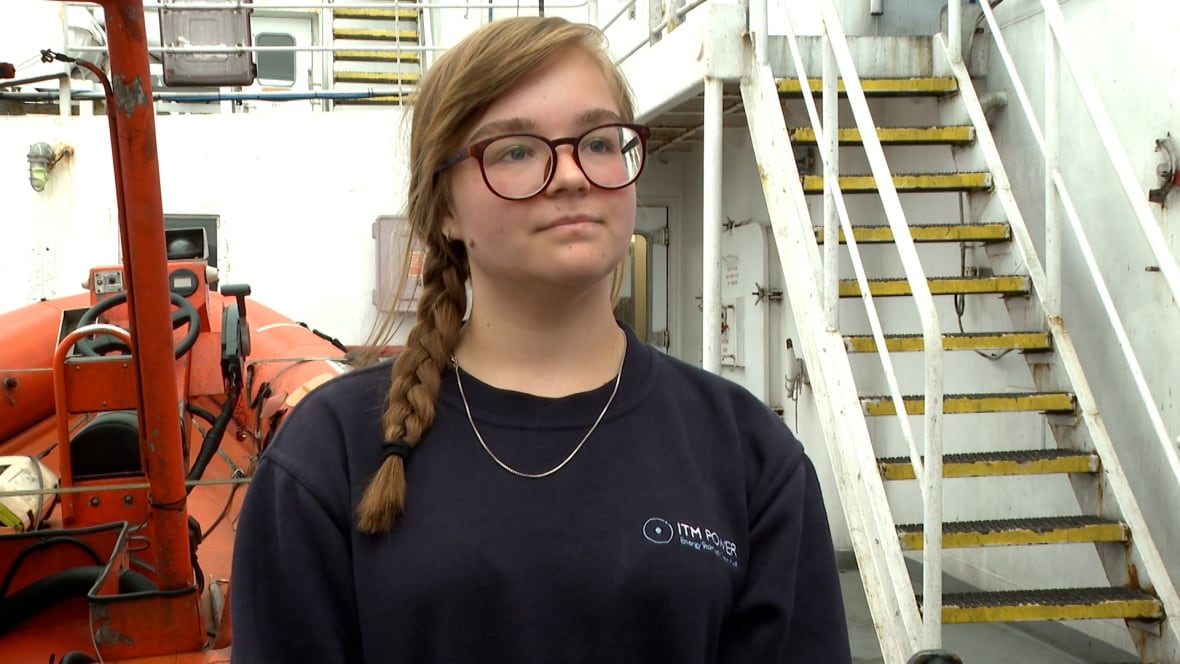 A girl with a braid in her hair and wearing glasses standing on the dock of a big vessel.