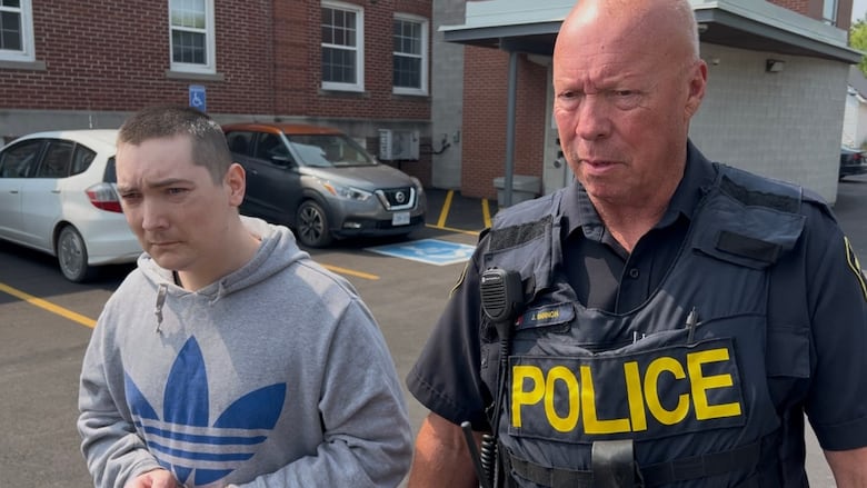 A man with buzzed dark hair and wearing a grey hoodie is escorted out of a brick building by an OPP officer.