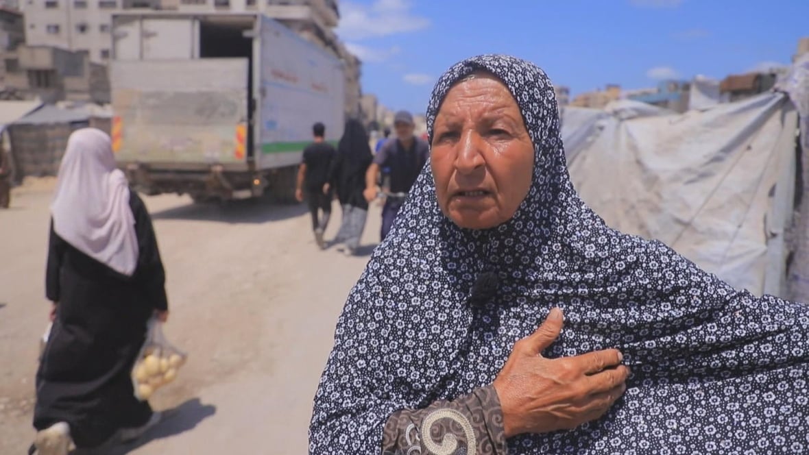 A woman wearing a blue floral headscarf gestures as she talks while standing outside a tent next to a dusty road.