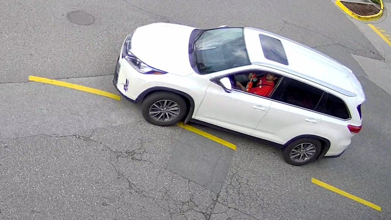 A CCTV image of a white SUV with a person with a red shirt behind the wheel and holding a red and white object out the window.