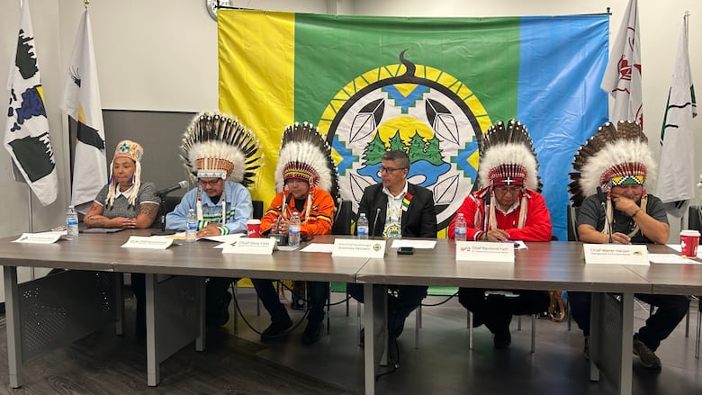 People sit at a table, many wearing feathered headdresses, with a large yellow, green, blue and white flag stretched out behind them. 
